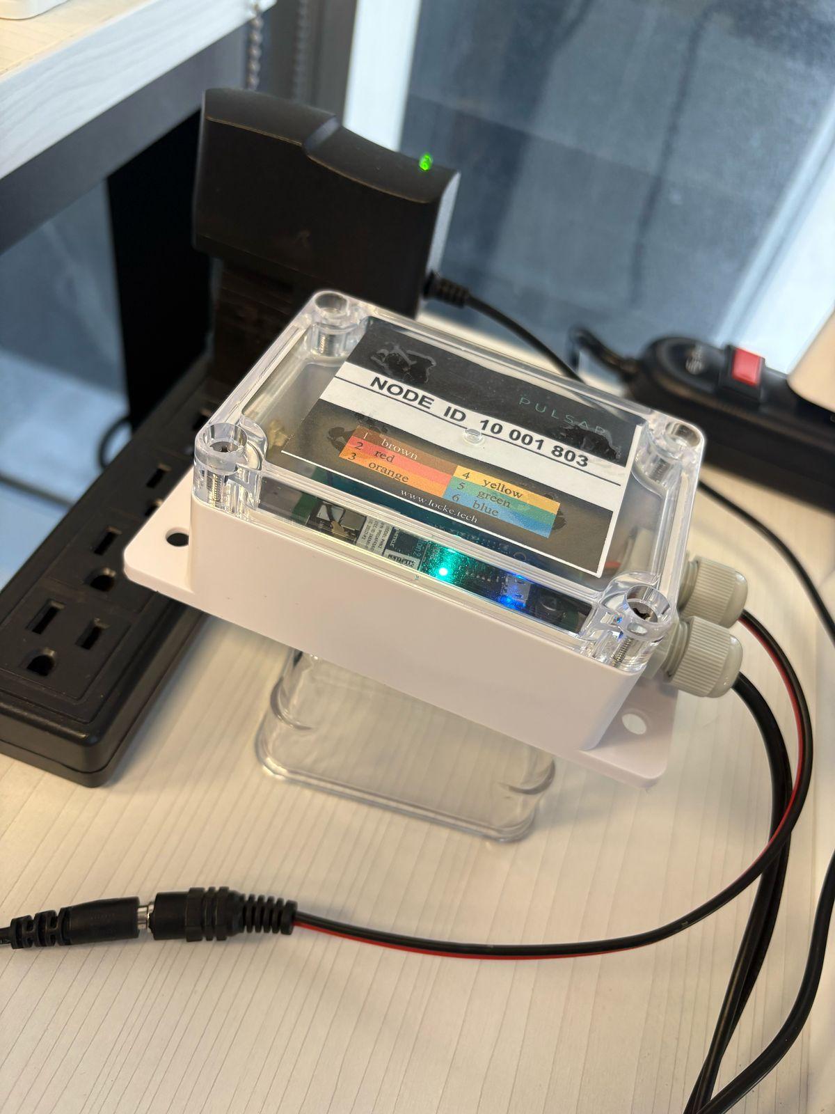 A Pulsar Smart Hub, a white box with a clear lid and a Node ID label, plugged into its power adapter, with the adapter's green LED and the hub's blue LED on.
