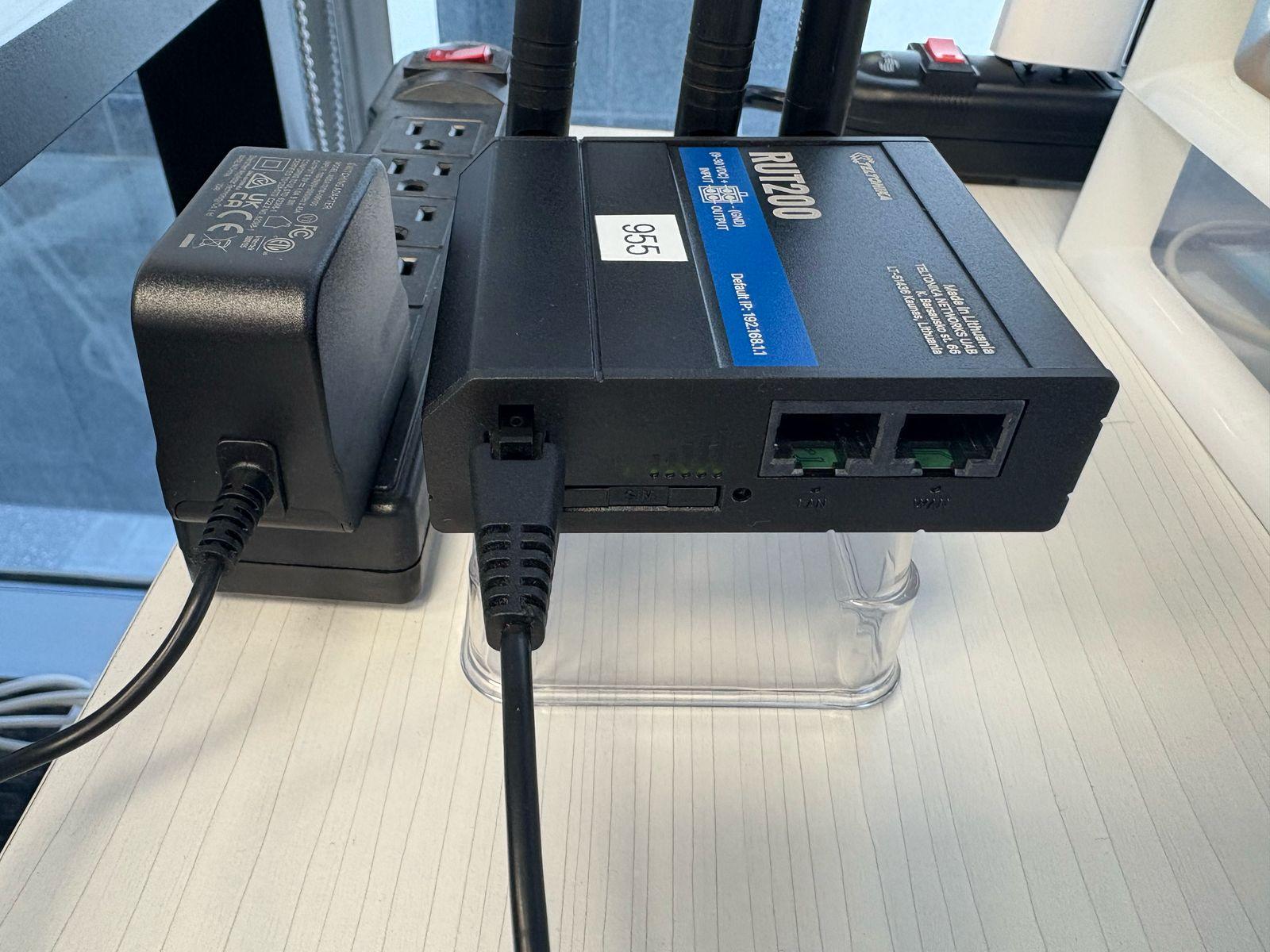 The router, a black Teltonika RUT200 with antennas, plugged into its power adapter, showing its status LEDs and LAN and WAN ports.